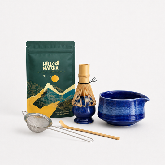 A matcha tea preparation set on a clean white background, featuring a green and gold Hello Matcha pouch, a blue ceramic chashaku, a bamboo whisk, a metal strainer, and a bamboo scoop.