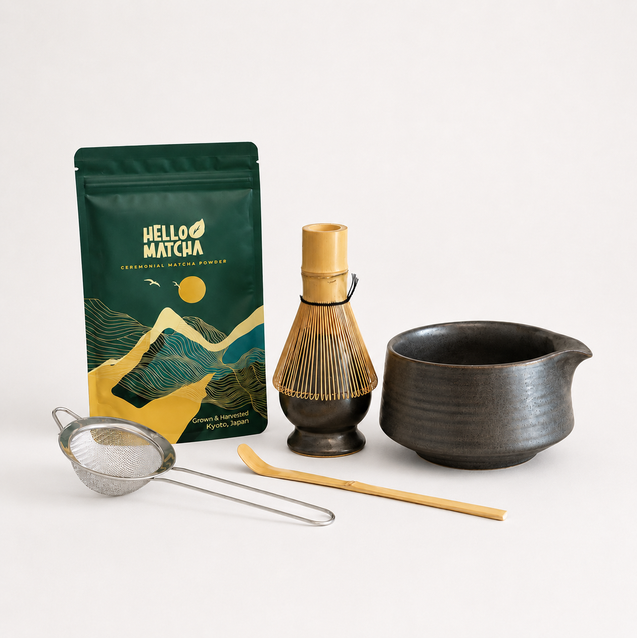 A matcha tea kit on a clean white background, featuring a green and gold Hello Matcha pouch, a bamboo whisk, a dark ceramic bowl, a metal strainer, and a bamboo scoop.