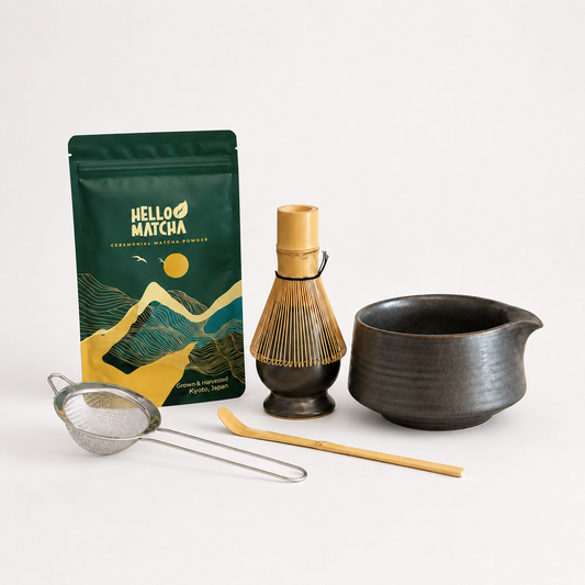 A matcha tea kit on a clean white background, featuring a green and gold Hello Matcha pouch, a bamboo whisk, a dark ceramic bowl, a metal strainer, and a bamboo scoop.