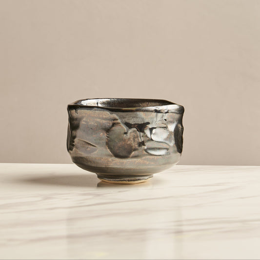 A handmade ceramic tea bowl with a textured, organic metallic finish in silver, bronze, and dark gray tones, sitting on a light marble surface against a neutral beige background.