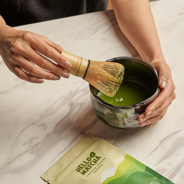 A person holds a bamboo whisk over a dark bowl filled with bright green matcha tea on a light marble surface. A package of Hello Muncha matcha is visible in the lower foreground.