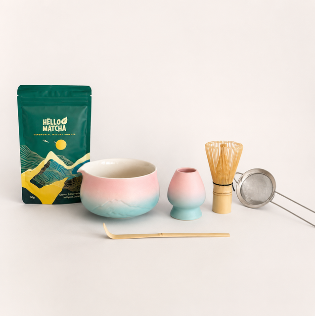 A matcha tea set arrangement on a clean white background, featuring a green Hello Matcha pouch, a pink and blue gradient ceramic bowl, a matching pink and blue gradient small vase, a bamboo whisk, a metal tea strainer, and a bamboo scoop.