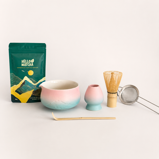 A matcha tea set arrangement on a clean white background, featuring a green Hello Matcha pouch, a pink and blue gradient ceramic bowl, a matching pink and blue gradient small vase, a bamboo whisk, a metal tea strainer, and a bamboo scoop.